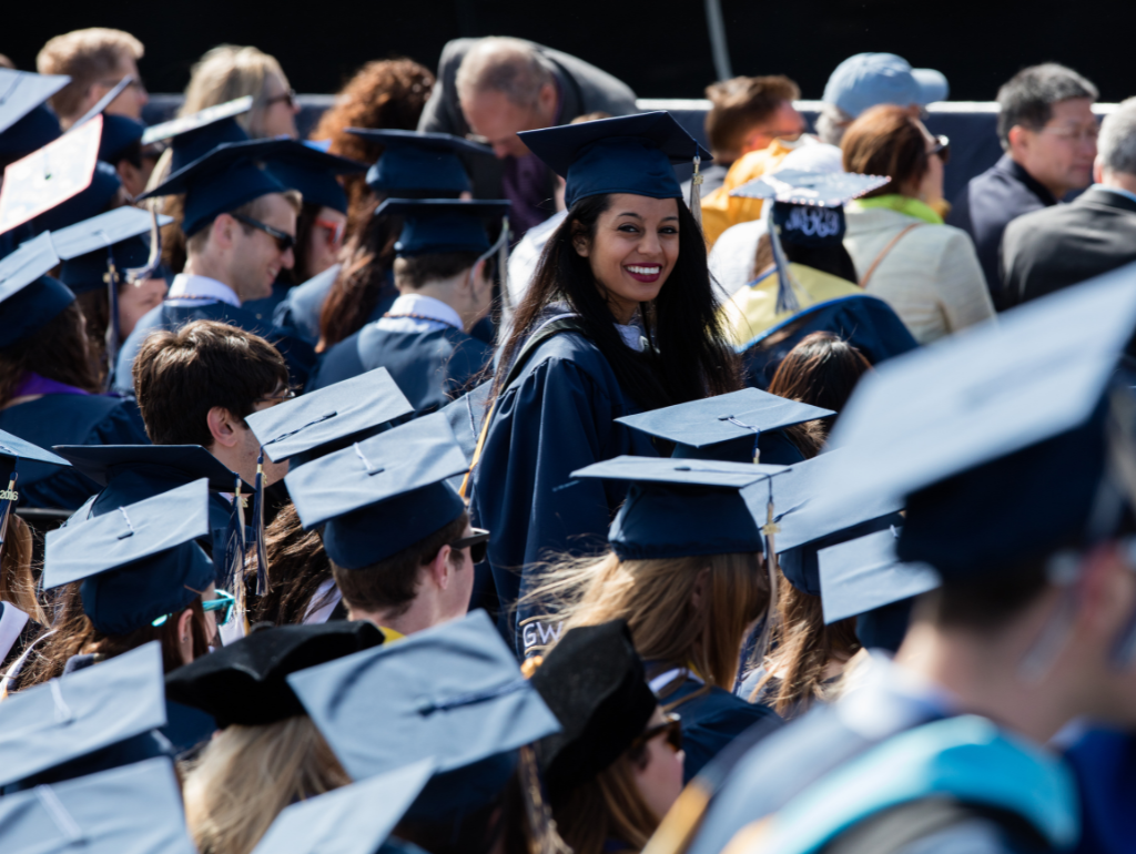 A graduating student smiling in a large crowd of fellow graduates wearing caps and gowns