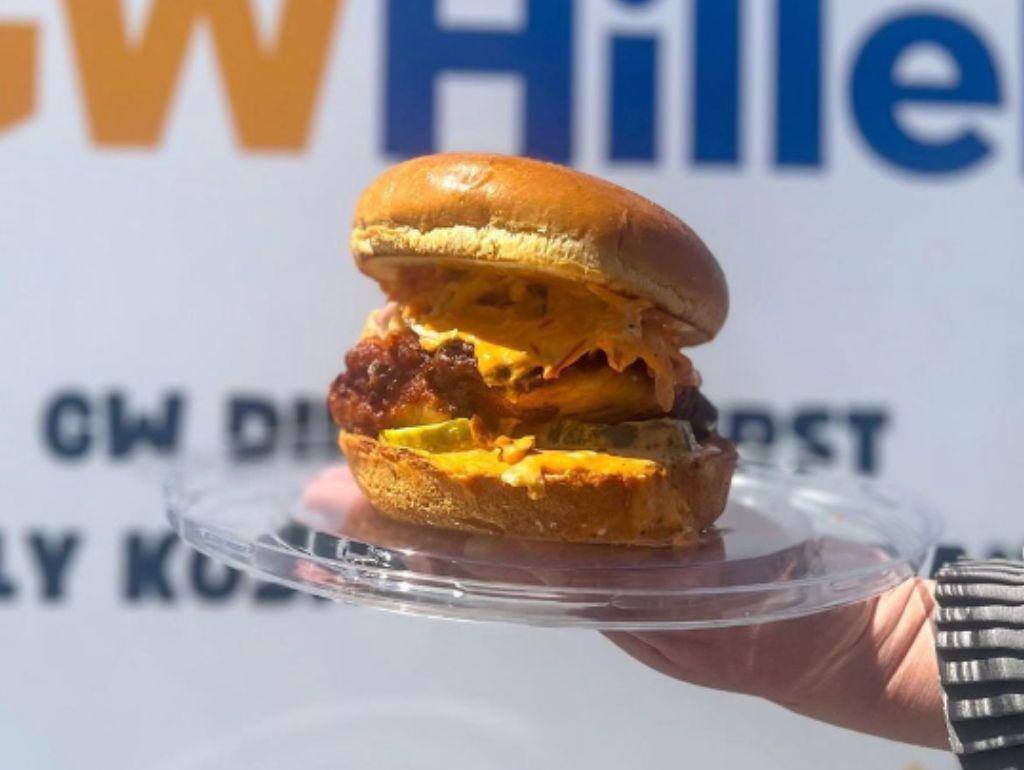 A fried chicken sandwich from GW Hillel held in front of a GW Hillel banner