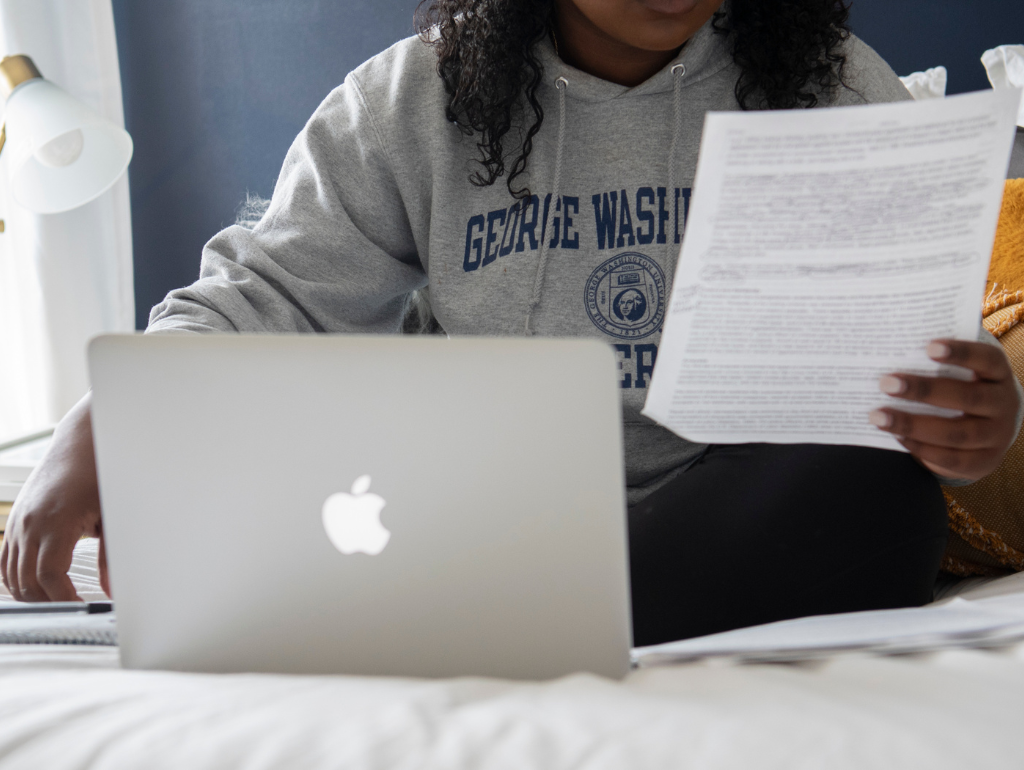 Student sitting on a bed with an open laptop examining a printed document