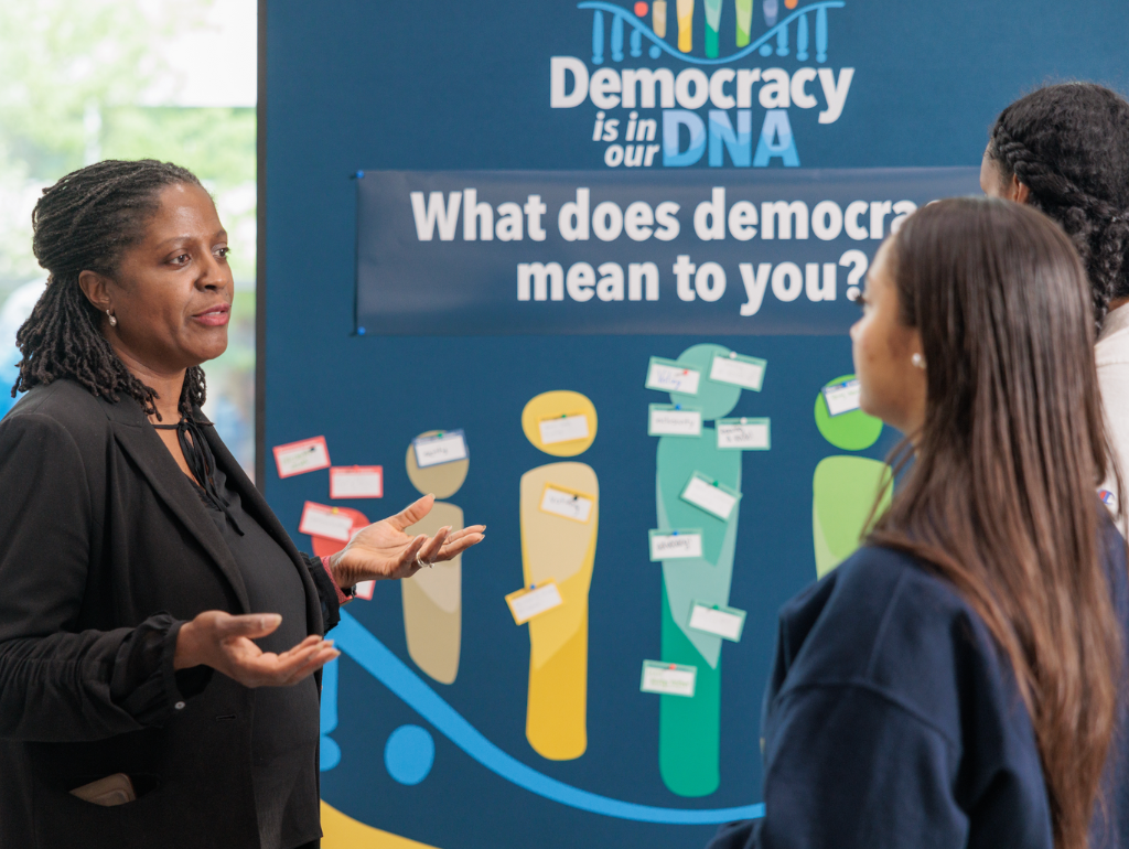 Vice Provost for Student Affairs and Dean of Students Colette Coleman talking with two students in front of a display board with the question "What does democracy mean to you?"
