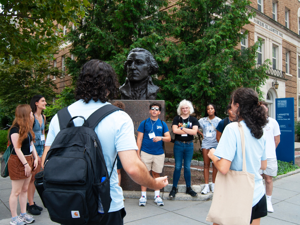 Group of students standing in a circle outside a GW residence hall with a bust statue of George Washington in the background
