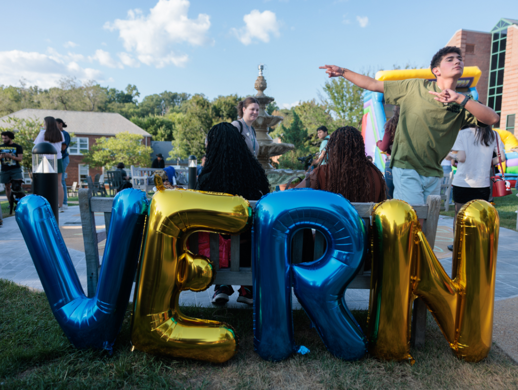 Blue and gold balloons spelling out "VERN" rest behind a bench on the MVC quad with student milling around at an event