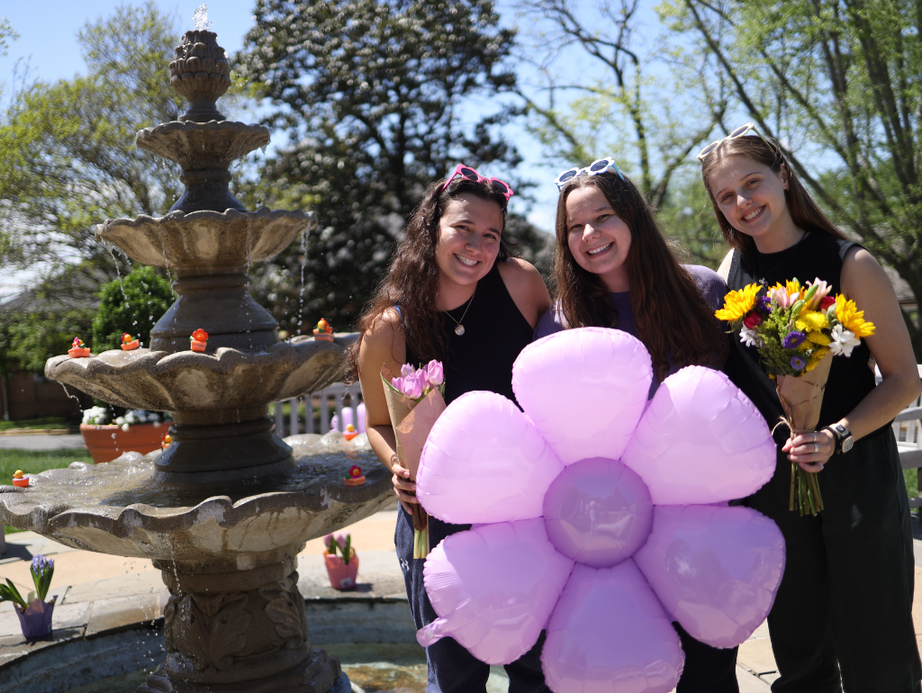 Three students posing in front of the Fountain with a flower made of purple balloons